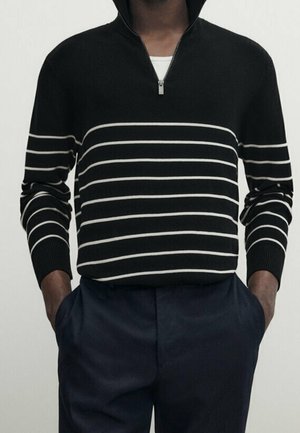 Homme portant un pull noir à fermeture éclair quart avec des rayures horizontales blanches et un pantalon foncé, mains dans les poches, sur fond uni.