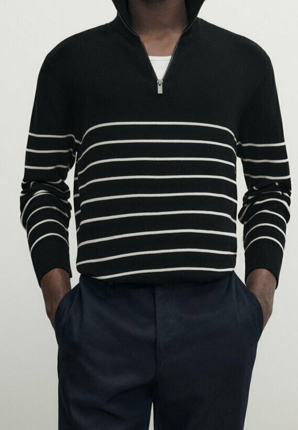 Homme portant un pull noir à fermeture éclair quart avec des rayures horizontales blanches et un pantalon foncé, mains dans les poches, sur fond uni.