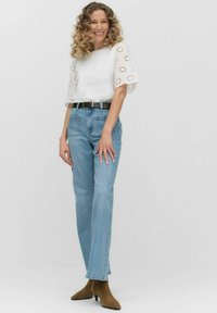 Vit topp med hålmönster, i kombination med ljusblå rakbenade jeans och bruna ankelskor. Neutral bakgrund framhäver outfitens egenskaper.