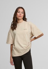 Beige overdimensjonert bomull T-skjorte med rund hals og korte ermer, med subtil tekstdetalj på forsiden.