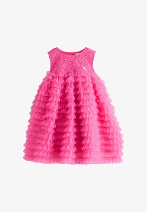 Niña joven con un vestido rosa brillante de volantes en capas y zapatos blancos de vestir con flores, de pie sobre un suelo claro con un fondo sencillo.