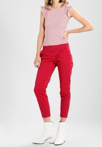 Red trousers with slim fit and cropped length, paired with a white top featuring red polka dots and ruffled sleeves. White ankle boots.