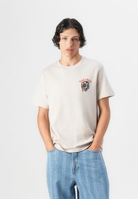 Beige katoenen t-shirt met een zwart en oranje hondenafbeelding op de linkerborst. Gecombineerd met lichtblauwe jeans met een kettingpatroon.