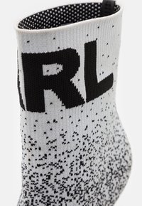 Gros plan sur une chaussette grise tricotée avec un motif pixelisé noir et de grandes lettres noires partiellement visibles près du bord-côte.