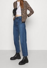 Žena v vzorovaném béžovém a černém saku, bílém crop topu, modrých džínách s vysokým pasem, černých platformových botách a černé crossbody tašce.