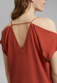 Top de color óxido hecho de una tela suave, con mangas cortas y un respaldo en forma de V con un detalle de tirante, que crea un look de hombros al descubierto.