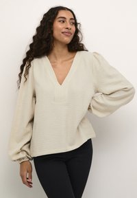 Beige V-Ausschnitt-Bluse aus strukturiertem Stoff, mit Puffärmeln und lockerer Passform, kombiniert mit dunklen Jeans. Schlichtes Design, keine sichtbaren Muster oder Akzente.