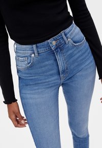 Jeans de talle alto azul claro con una textura suave, cinco bolsillos y un desvanecido sutil, combinados con una blusa ajustada de manga larga negra.