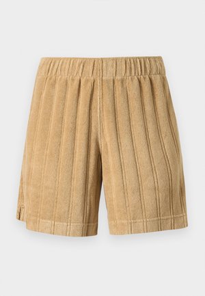 Homme portant une casquette beige, un t-shirt blanc avec logo, un short moutarde texturé et des sandales marron foncé slip-on, debout devant un fond uni.