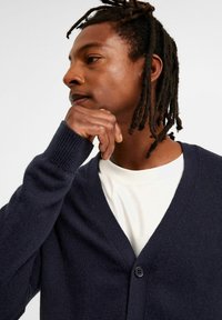 Marinefarbener Strickcardigan mit V-Ausschnitt, ausgestattet mit einem einzigen Knopfverschluss und gerippten Bündchen. Über einem weißen Hemd getragen.