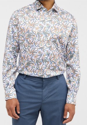 Homme portant une chemise blanche à manches longues avec un motif cachemire multicolore rentrée dans un pantalon habillé bleu, debout les bras détendus.
