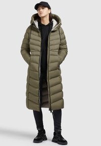Lång olivgrön pufferjacka med horisontell quiltning, huva, dragkedja fram och sidofickor. Bärs över en svart hoodie och mörka jeans.