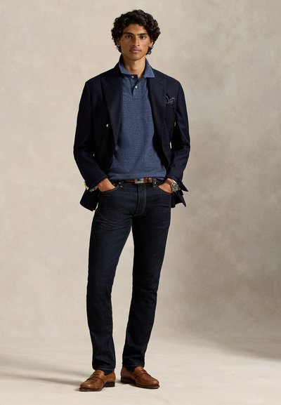 Homme aux cheveux bouclés et foncés, debout les mains dans les poches, portant un blazer bleu marine, un polo bleu, un jean foncé, une ceinture marron, une montre et des chaussures en cuir marron.