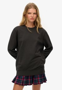 Mujer joven con cabello rubio largo, vistiendo un sudadera negra de gran tamaño y una falda plisada a cuadros rojos y azules, de pie con las manos detrás de la espalda.