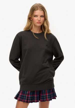 Jonge vrouw met lang blond haar, gekleed in een zwarte oversized sweatshirt en een geplooide rok in rood en blauw ruitpatroon, staande met handen achter haar rug.