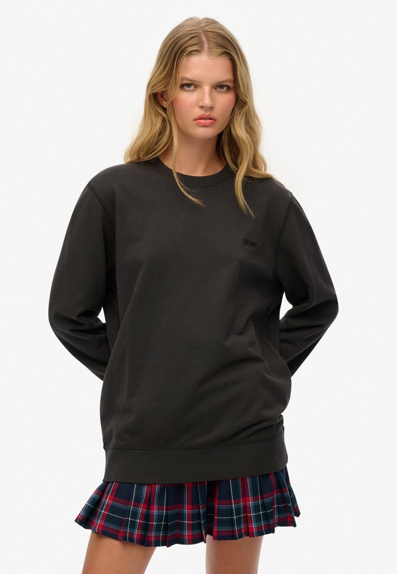 Mujer joven con cabello rubio largo, vistiendo un sudadera negra de gran tamaño y una falda plisada a cuadros rojos y azules, de pie con las manos detrás de la espalda.