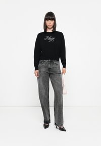 Sudadera negra con el logotipo "Hugo" bordado, combinada con unos vaqueros de pierna ancha gris de talle alto. Tacones negros de punta y un pequeño bolso rosa.