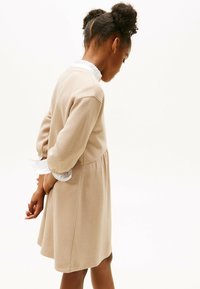 Robe beige côtelée avec un corsage ajusté, une jupe évasée et des manches élastiques. Présente un col en dentelle blanc et des détails aux poignets.