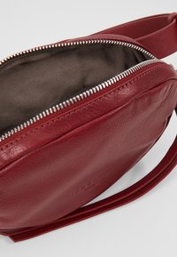 Borsa a tracolla rossa in pelle con forma rotonda, chiusura con zip e fodera in tessuto marrone. Presenta una lunga tracolla regolabile e un logo impresso.