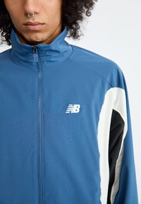 Ungdom med krøllete hår som har på seg en blå glidelåsjakke fra New Balance med hvite og svarte detaljer på ermet.