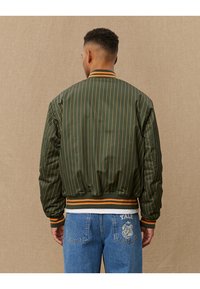 Grüne Bomberjacke mit vertikalen orangefarbenen Streifen, geripptem Kragen und Bündchen. Mit einem Gummibund in Kombination mit einem weißen Streifen und glatter Stoffstruktur.