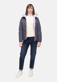 Giacca imbottita viola con fodera lavanda a contrasto, chiusura con zip e cappuccio attaccato, abbinata a jeans scuri e sneakers bianche.