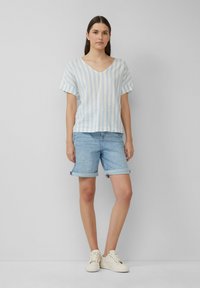 Jasnoniebieska i biała, w paski, krótka bluzka z dekoltem w serek, sparowana z jasnymi dżinsowymi szortami. Modelka ma na sobie kremowe sneakersy i stoi na tle w neutralnych kolorach.