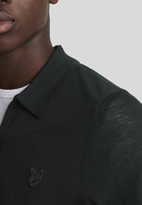 Schwarzes gestricktes Polohemd mit einem gerippten Kragen und einem kleinen gestickten Emblem auf der linken Seite. Strukturierter Stoff, über einem weißen Hemd getragen.