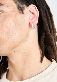 Zilveren hoopoorbel met een gestructureerd oppervlak en kleine accentstenen, gedragen in een oor met dreadlocks; de huid vertoont lichte sproeten.
