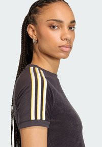 Čierne tričko s krátkym rukávom so žltými a bielymi prúžkami na ramenách, vyrobené z jemnej látky. Má okrúhly výstrih a rebrované manžety.