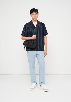 Giovane uomo con camicia a righe blu navy a maniche corte, jeans azzurri, sneakers bianche, che tiene una borsa a tracolla nera su una spalla.