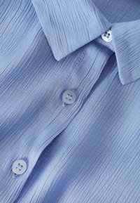 Light blue shirt made of textured fabric with a vertical rib pattern, featuring a fold-down collar and two blue buttons on the placket.