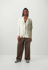 Cream-colored textured blazer with a collar and two buttons, paired with loose brown trousers and black-and-white sneakers.