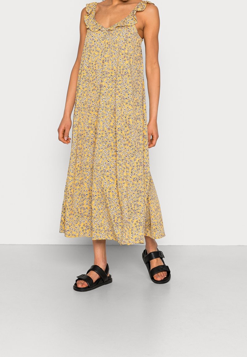 Femme portant une robe midi jaune à fleurs sans manches avec un col à volants et des sandales noires à bout ouvert avec des brides larges sur un sol gris.
