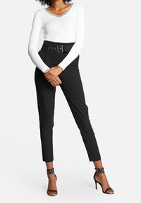 Mujer vestida con pantalones negros de cintura alta con cinturón, camiseta blanca de manga larga con escote en V y sandalias de tacón alto negras con tachuelas, de pie con las manos entrelazadas.