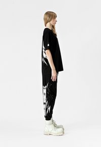 Camiseta negra oversized con diseño gráfico blanco en la parte trasera, combinada con pantalones negros con detalles impresos y botas gruesas de color claro.