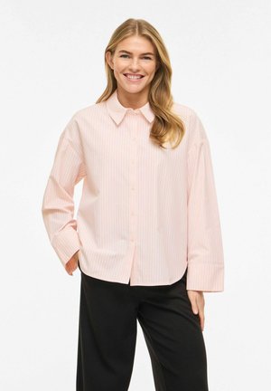 Smilende kvinne med langt blondt hår iført en lys rosa stripete skjorte med knapper og svarte bukser, stående med den ene hånden i lomma.