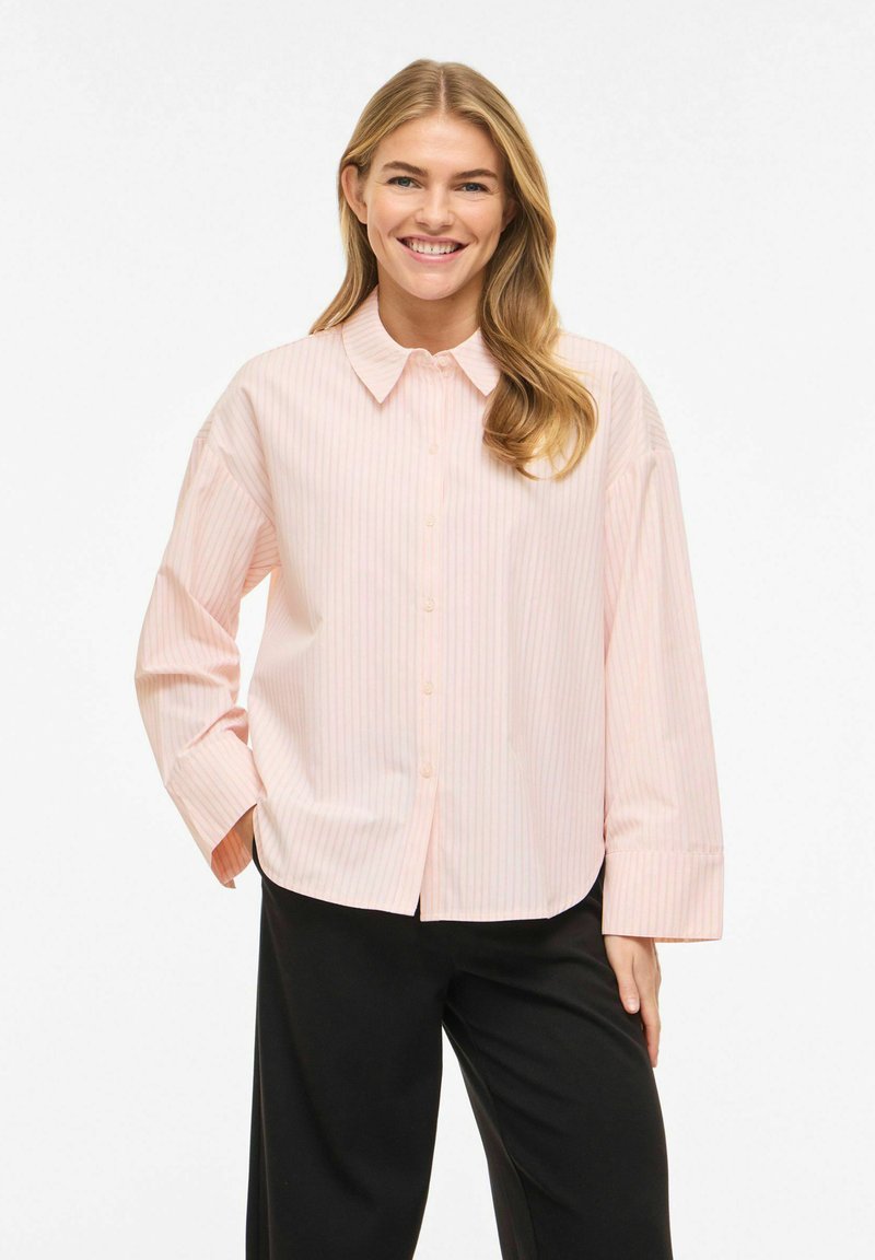 Femme souriante aux longs cheveux blonds, portant une chemise à rayures rose clair boutonnée et un pantalon noir, debout avec une main dans la poche.