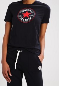 Osoba v černém tričku Converse All Star s červenou hvězdou a černých kalhotách s bílým šnůrkovým pasem a logem na kapse.