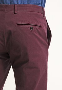 Pantaloni bordeaux con una texture liscia, presentano una tasca posteriore con chiusura a bottone e un sottile logo, abbinati a una camicia blu.