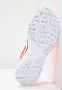 Sole of a white athletic shoe with geometric tread pattern, red fabric upper, leaning against a white corner wall.