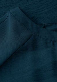 Türkises Gewebe mit glatter Textur, das über einen runden Ausschnitt und sich kreuzende Nahtdetails verfügt. Das Material scheint leicht und fällt sanft.