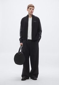 Zwarte jas met ritssluiting en gebreide textuur met logo, gecombineerd met losse zwarte broek. Houdt een ronde zwarte tas met handvat en gouden accent vast.