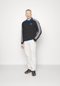 Sweatshirt noir avec des rayures blanches sur les manches, arborant un petit logo, associé à un pantalon blanc et des baskets noires avec des accents orange.