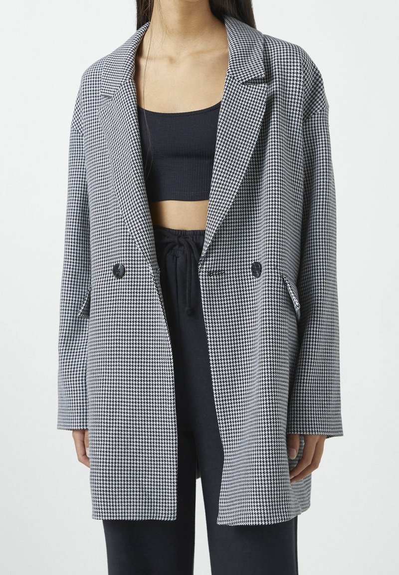 Blazer a cuadros en blanco y negro, con un corte holgado, solapas con muesca, cierre de doble botón y bolsillos laterales. Textura de tela suave.