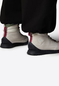 Bottes de randonnée beiges avec semelles foncées ; présentant un accent bordeaux au talon, des œillets en métal et une surface en cuir texturée.