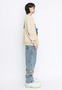 Sudadera beige con un gráfico de oso texturizado, combinada con jeans azul claro que tienen un parche de oso en la rodilla. Zapatillas de plataforma blancas y voluminosas.