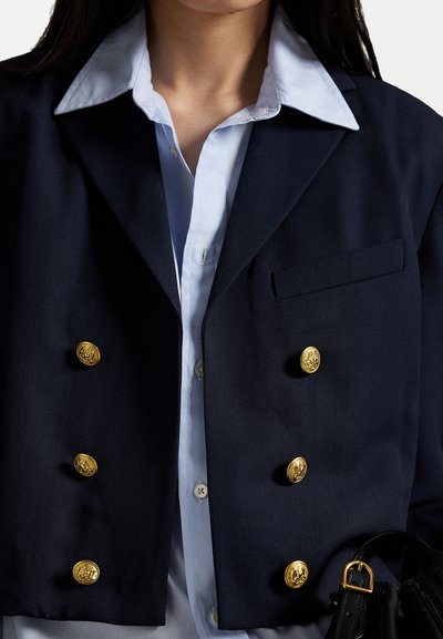 Navy double-breasted blazer with gold buttons, broad lapels, and a chest pocket, worn over a light blue collared shirt. Black bag visible.