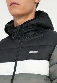 Sort og grå boblejakke med hætte. Har en hvid vandret stribe og logopatch. Glat tekstur med quiltede sektioner.