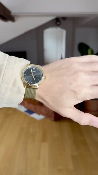 Kuldtooni kell musta näo, hõbedast metallvõrgust rihmaga, minimalistlik disain, ruudukujulised tunni markerid ja logo kell 12 asendis.